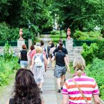 Eine Gruppe von Menschen geht auf einem von Bäumen gesäumten Weg in einem Park spazieren, im Hintergrund sind Stein- und Eisengeländer zu sehen.