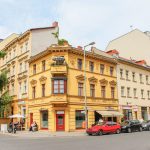 Ein gelbes Eckgebäude an einer Stadtstraße mit geparkten Autos und Passanten in der Nähe.