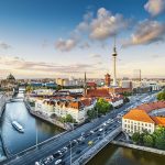 Eine Luftaufnahme fängt die lebendige Stadtlandschaft ein, wo sich ein gewundener Fluss an dem berühmten Fernsehturm und historischen Gebäuden vorbeischlängelt, alles unter einem blauen Himmel mit Wolken. Genau wie ein Team in Harmonie zusammenarbeitet, erschaffen diese Elemente ein atemberaubendes urbanes Bild.