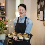 Person in einer Schürze hält ein Tablett mit Desserts, im Hintergrund sind Blumen und eine Karte in einem Café zu sehen.