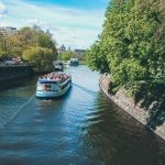 An einem sonnigen Tag fährt ein Boot mit Passagieren einen von Bäumen gesäumten Kanal entlang.