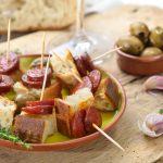 Ein Teller Tapas mit Brot, Oliven und Chorizo-Spießen auf einem rustikalen Holztisch.