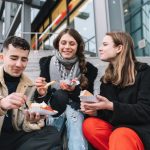 Drei Menschen sitzen auf Stufen, lächeln und essen Straßenessen von Papptabletts.