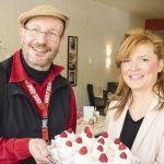 Zwei Menschen lächeln und halten in einem gemütlichen Raum einen mit Schlagsahne und Himbeeren garnierten Kuchen.