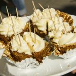 Sechs Stücke Karottenkuchen mit Frischkäse-Zuckerguss und Zahnstocher-Spießen auf einem weißen Teller.