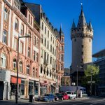 Straßenansicht einer Stadt mit historischen Gebäuden und einem hohen Steinturm unter einem klaren blauen Himmel.