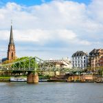 Malerische Aussicht auf einen Fluss, eine Brücke und einen Kirchturm mit historischen Gebäuden und einem bewölkten Himmel.