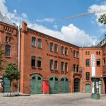 Historisches rotes Backsteingebäude mit grünen Türen und einem Innenhof unter einem blauen Himmel mit Wolken.