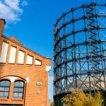 Altes rotes Backsteingebäude neben einem großen Gasbehälter aus Metall vor einem strahlend blauen Himmel.