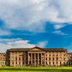 Ein großes, historisches Gebäude mit Säulen unter einem teilweise bewölkten blauen Himmel, umgeben von einer Rasenfläche und Bäumen.