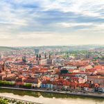 Luftaufnahme einer europäischen Stadt mit Gebäuden mit roten Dächern und einem Fluss unter einem teilweise bewölkten Himmel.