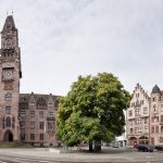 Historische Steingebäude und ein Baum auf einem offenen Platz an einem bewölkten Tag.