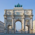 Triumphbogen mit Statuen darauf, eingerahmt von historischen Gebäuden unter einem klaren blauen Himmel.