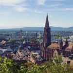 Luftaufnahme einer Stadt mit einem Kathedralenturm, umgeben von Grün und fernen Hügeln unter einem blauen Himmel.