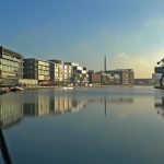 Blick auf den Kanal mit modernen Gebäuden, einem großen Kran und einem klaren blauen Himmel, der sich in der ruhigen Wasseroberfläche spiegelt.