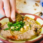 Eine Hand tunkt Falafel in cremigen Hummus, garniert mit Petersilie und Kichererbsen, im Hintergrund ist Brot zu sehen.