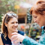 Zwei Frauen lächeln und unterhalten sich draußen, umgeben von Grün. Eine Frau hält eine Wasserflasche.