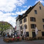 Ein europäischer Platz mit Cafétischen im Freien, spazierenden Menschen und historischen Gebäuden unter einem teilweise bewölkten Himmel.