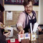 Ein Barista in Schürze bedient einen Kunden, der mit Stäbchen an der Theke eines rustikalen Cafés isst.