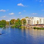 Flussszene mit einem Brunnen, umgeben von Gebäuden und Bäumen unter einem klaren blauen Himmel.