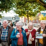 Vier ältere Freunde lachen und halten Kaffeetassen und eine Karte in den Händen, während sie auf einer sonnigen, von Bäumen gesäumten Straße spazieren gehen.