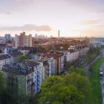 Panoramablick auf eine Stadtlandschaft mit Bäumen, Gebäuden, einem Fluss und einer Brücke unter einem teilweise bewölkten Himmel bei Sonnenuntergang.