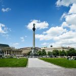 Eine malerische Aussicht auf einen Stadtplatz mit einer zentralen Säule, umgeben von Gebäuden und grünen Rasenflächen unter einem teilweise bewölkten Himmel.