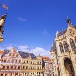 Historischer europäischer Platz mit reich verzierten mittelalterlichen Gebäuden und einer Statue unter einem klaren blauen Himmel.