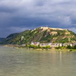 Panoramablick auf einen Fluss mit einer Festung auf einem Hügel, Booten und üppigem Grün unter einem bewölkten Himmel.