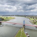 Luftaufnahme einer roten Hängebrücke über einen breiten Fluss inmitten grüner Landschaften und Industriegebiete unter bewölktem Himmel.