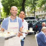 Zwei lächelnde Kellner tragen Tabletts mit Desserts auf einer sonnigen Straße im Freien, im Hintergrund sitzende Menschen.