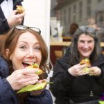 Zwei Menschen lächeln und essen Sandwiches, während sie vor einem Café sitzen.