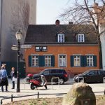 Straßenszene mit einer Gruppe von Menschen, geparkten Autos, einem Motorrad und historischen Gebäuden im Hintergrund.