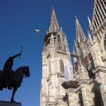 Statue eines Ritters zu Pferd, die sich vor einer gotischen Kathedrale mit hohen Türmen unter einem klaren blauen Himmel abhebt.