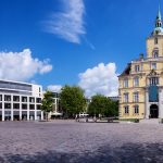 Ein sonniger Stadtplatz mit einem historischen gelben Gebäude und einem modernen weißen Gebäude unter einem blauen Himmel mit Wolken.