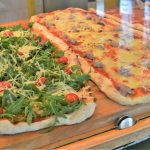 Zwei rechteckige Pizzen: eine mit Rucola und Kirschtomaten, die andere mit Käse und Fleisch, auf einem Holzbrett.