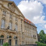 Historisches Gebäude mit reich verzierter Fassade unter teilweise bewölktem Himmel, umgeben von Grün.