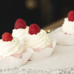 Vier Cupcakes mit Schlagsahne und Himbeeren auf einem Tablett mit Spitzenpapier.