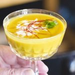 Eine Hand hält ein Glas cremige, goldene Suppe, garniert mit Sahne, Koriander und dünnen roten Fäden.