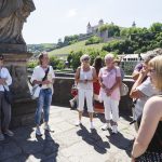 Eine Gruppe von Menschen steht auf einer sonnigen Brücke in Würzburg vor einer malerischen Kulisse aus Hügeln und historischen Gebäuden.