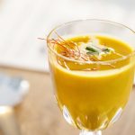 Ein Glas gelbe Suppe, garniert mit Kräutern und roten Strängen auf einem Holztisch.