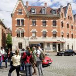 An einem sonnigen Tag steht eine Gruppe von Menschen in der Nähe eines historischen Backsteingebäudes, davor parken Autos.