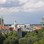 Luftaufnahme einer Stadt mit einem historischen Gebäude, einer grünen Kuppel, modernen Wolkenkratzern und üppigem Grün unter einem bewölkten Himmel.