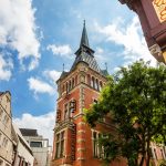 Historischer Backsteinturm mit Spitzdach vor einem blauen Himmel mit Wolken, eingerahmt von nahe gelegenen Gebäuden und einem Baum.