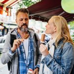 Ein Mann und eine Frau genießen ein Eis, während sie eine Stadtstraße entlanggehen, umgeben von parkenden Autos und Grünflächen.