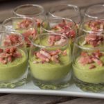 Kleine Gläser gefüllt mit grüner Mousse, garniert mit geriebenem Speck, angerichtet auf einem weißen Tablett.