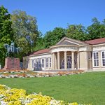 Ein historisches Gebäude mit einem Portikus und einer Reiterstatue, umgeben von leuchtend grünem Laub und Blumen.