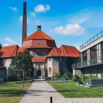 Historisches Gebäude mit rotem Dach, angrenzendem modernen Gebäude und einem Kopfsteinpflasterweg in einer Rasenfläche.