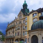 Historisches Gebäude mit einer grünen Kupferkuppel vor einem bewölkten Himmel, umgeben von Bäumen und Ladenfronten.