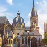 Historische Kathedrale mit komplexer Architektur, Turmspitze und Kuppel, umgeben von Bäumen unter einem strahlend blauen Himmel.